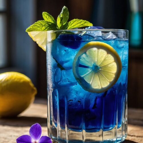 Glass of blue lemonade with mint leaves and lemon slice on a wooden table, surrounded by fresh lemons and butterfly pea flowers.